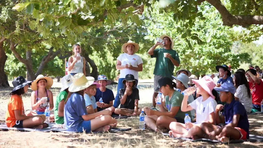 Groupe d’enfants et d’animateurs en périscolaire se reposant à l’ombre sous les arbres pendant une forte chaleur, en train de boire de l’eau et de se protéger du soleil